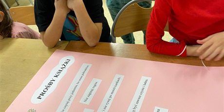 Powiększ grafikę: Zdjęcie przedstawia 2 dzieci, które stoją przy stoliku. Na stole leży różowy karton z kartkami, na których są zapisane zdania - prośby książki: Nie czytaj przy jedzeniu./ Nie pisz i nie rysuj po mnie.