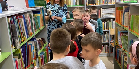 Powiększ grafikę: Zdjęcie przedstawia grupę dzieci stojącą między regałami książek. Na końcu grupy stoi pani, która pokazuje dzieciom książkę.