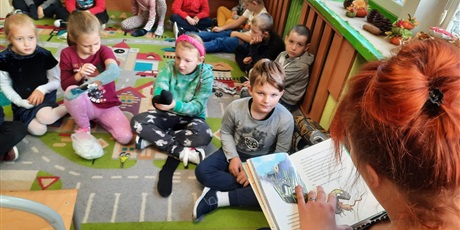Powiększ grafikę: Na zdjęciu pani czyta książkę dzieciom, które siedzą na dywanie.