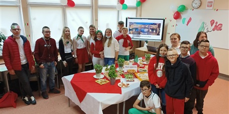Powiększ grafikę: Obraz przedstawia uczniów i wychowawcę stojących wokół stołu z daniami kuchni włoskiej. W tle widoczny jest obraz budowli wyświetlanej na tablicy interaktywnej oraz kolorowy napis Italia.
