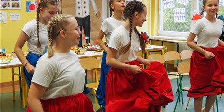 Powiększ grafikę: Obraz przedstawia pięć tańczących uczennic. Wszystkie są ubrane w białe koszulki i mają zaplecione dwa warkocze. Trzy z nich mają czerwone spódnice, a dwie niebieskie. W tle widoczne są francuskie potrawy oraz rysunek przedstawiający kucharza.