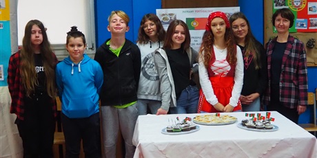 Powiększ grafikę: Obraz przedstawia siedmiu uczniów i nauczycielkę pozujących za stołem, na którym stoją babeczki z flagami Portugalii. W tle widać flagę Portugalii oraz zdjęcia potraw kuchni portugalskiej.