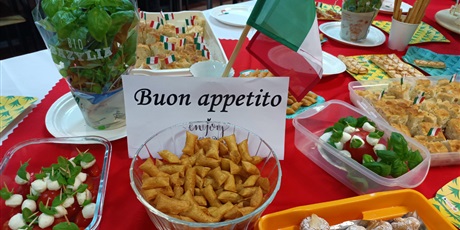 Powiększ grafikę: Obraz przedstawia stół z różnymi potrawami kuchni włoskiej (ciasteczka, ciasto pokrojone na kawałki z wbitymi wykałaczkami z flagą Włoch, koreczki z mozzarelli, pomidorków i bazylii, paluszki Grissini). Na środku stoi flaga Włoch oraz napis Buon appetito.
