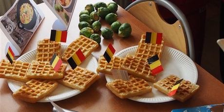 Powiększ grafikę: Obraz przedstawia gofry ułożone na dwóch talerzach, w które powbijane są flagi Belgii. W tle widoczne są dwa zdjęcia przedstawiające potrawy, czekolada i brukselki.