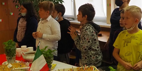 Powiększ grafikę: Obraz przedstawia uczniów i nauczycielkę stojących za stołem z włoskimi potrawami. Wszyscy patrzą w lewą stronę.