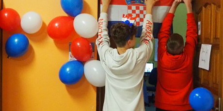 Powiększ grafikę: Obraz przedstawia dwóch uczniów stojących tyłem i wieszających flagę Chorwacji nad drzwiami.