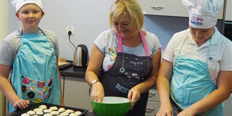 Powiększ grafikę: Obraz przedstawia dwóch uczniów i nauczycielkę w fartuchach kuchennych (uczniowie mają na głowie czapki kucharza). Nauczycielka trzyma dłoń w zielonej misce, a przed uczniami widoczne są małe ciastka ułożone na czarnej blaszce do pieczenia.