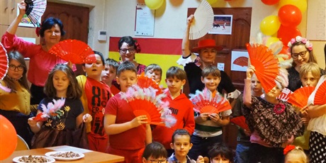 Powiększ grafikę: Obraz przedstawia wielu uczniów i cztery nauczycielki, trzymających hiszpańskie wachlarze. W tle widoczna jest flaga Hiszpanii i balony.