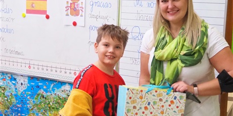Powiększ grafikę: Obraz przedstawia uśmiechniętą nauczycielkę wręczającą uczniowi prezent (kolorowa torebka).