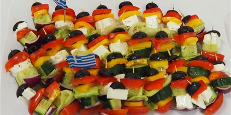 Powiększ grafikę: Obraz przedstawia koreczki warzywno- serowe ułożone na talerzu. Pomiędzy koreczkami włożone są flagi Grecji.