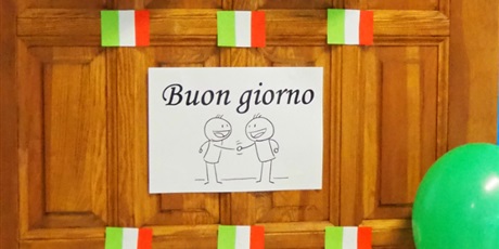 Powiększ grafikę: Obraz przestawia drzwi z naklejoną kartką z napisem Buon giorno i z ikonką dwóch podających sobie ręce postaci. Na drzwiach widoczne są także małe flagi Włoch, kod QR oraz balony.