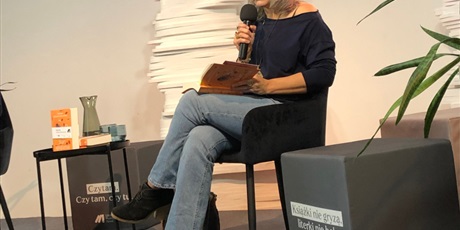 Powiększ grafikę: Zdjęcie przedstawia Roksanę Jędrzejewską-Wróbel, która siedzi na scenie z mikrofonem. W drugiej ręce trzyma książkę. Po lewej stronie stoi stolik.
