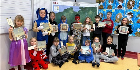 Powiększ grafikę: Grupa dzieci stoi i siedzi w sali lekcyjnej. W rękach trzymają książki. Za nimi wisi mapa Polski.