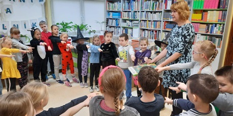 Powiększ grafikę: Na zdjęciu dzieci w bibliotece stoja w kręgu, trzymają wyciągnięte przed siebie prawe ręce i 2 palce.