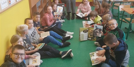 Powiększ grafikę: Zdjęcie przedstawia grupę dzieci, które siedzą na dywanie w kole i czytają książki. Pani siedzi na krześle również z książką.
