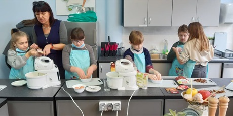 Powiększ grafikę: Zdjęcie przedstawia pięcioro dzieci i nauczycielką w kuchni szkolnej. Dzieci mają na sobie kuchenne fartuszki. Na stołach stoją miksery, miski. Dzieci przygotowują ciasto.
