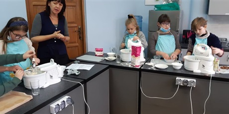 Powiększ grafikę: Zdjęcie przedstawia czworo dzieci i nauczycielkę w kuchni szkolnej. Uczniowie mają na sobie fartuszki kuchenne. Na stołach stoją miksery, mąka, deski do krojenia, szklanki. Dzieci dokładają składniki 