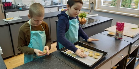 Powiększ grafikę: Zdjęcie przedstawia dwóch chłopców w kuchni szkolnej. Obaj mają na sobie fartuszki kuchenne. Na stole mają blaszkę do pieczenia, na której układają ciasto w formie okrągłych ciasteczek.
