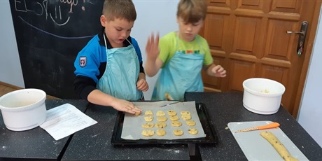 Powiększ grafikę: Zdjęcie przedstawia dwóch chłopców w kuchni szkolnej. Obaj mają na sobie fartuszki kuchenne. Na stole są miski, kartka, stolnica, blaszka do pieczenia i nóż. Chłopcy układają na blaszce ciasto w kszta