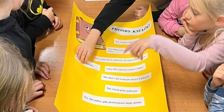 Powiększ grafikę: Zdjęcie przedstawia dzieci zebrane wokół żółtego kartonu. Przyklejają na niego zdania wypisane na kartkach.