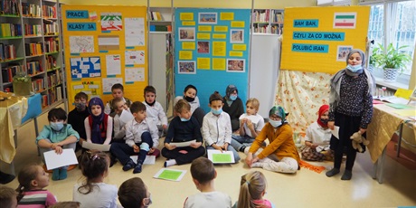 Powiększ grafikę: Zdjęcie przedstawia grupę dzieci w sali bibliotecznej. Wszyscy siedzą na okrągłych siedziskach na podłodze. Jedna dziewczynka stoi po prawej stronie, w ręku trzyma pluszowego misia. Naprzeciwko (tyłem