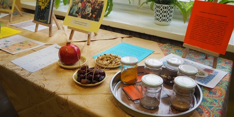 Powiększ grafikę: Zdjęcie przedstawia długi stół przykryty złotym obrusem. Na stole stoją małe sztalugi z książką i zdjęciami, talerzyki z daktylami, pistacjami i granatem, kartki, taca ze słoiczkami. W tle okno i para